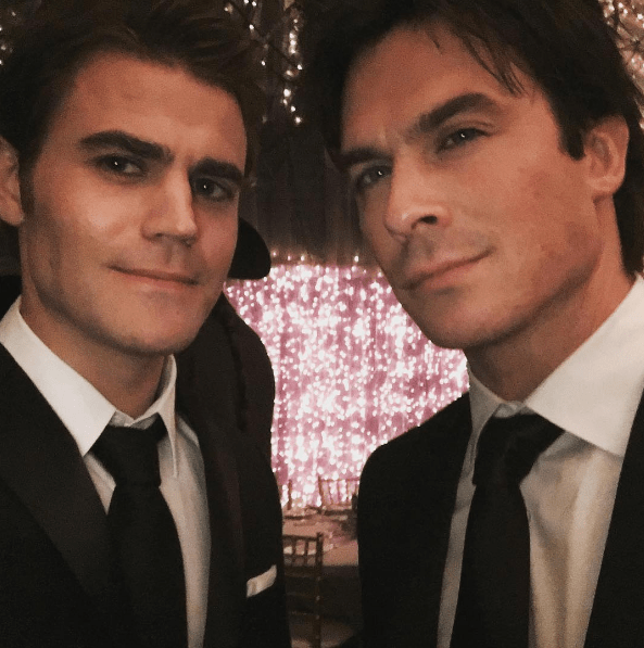 Selfie de Paul Wesley e Ian Somerhalder; os atores usam ternos pretos com camisa social branca e gravata preta, eles sorriem levemente para câmera