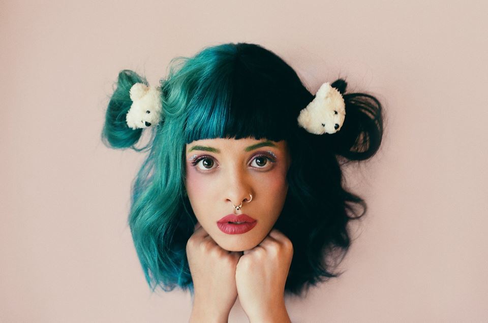 Melanie Martinez posando para foto com as duas mãos apoiadas no queixo; metade de seu cabelo em tons de verde e azul, outra metade preto