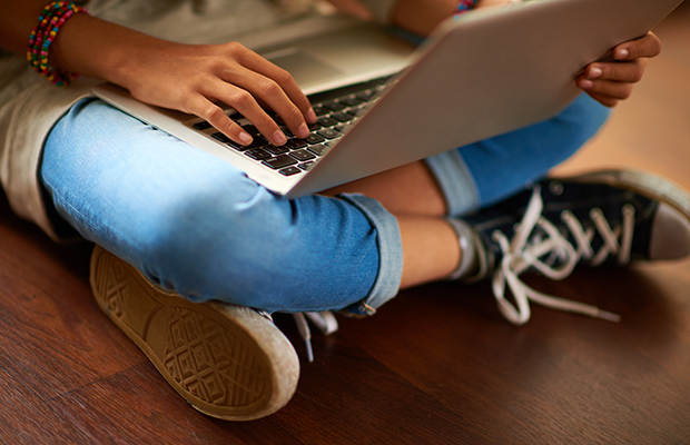 A imagem mostra uma pessoa sentada. É possível ver apenas suas pernas cruzadas, usando uma calça jeans e tênis All Star preto, enquanto apoia um notebook no colo. É possível ver suas mãos, com pulseiras coloridas, mexendo no aparelho. 