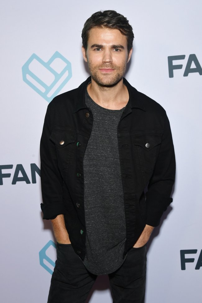 Paul Wesley posando para foto no carpet da Comic Con em 2018, ele usa camiseta, calça e jaqueta de cor preta e sorri levemente