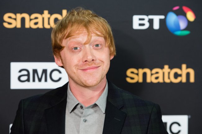 “Snatch” TV Show Premiere – Red Carpet Arrivals Rupert Grint em carpet de premiere sorrindo levemente para a câmera e arregalando os olhos; ele usa uma camisa cinza com blazer escuro