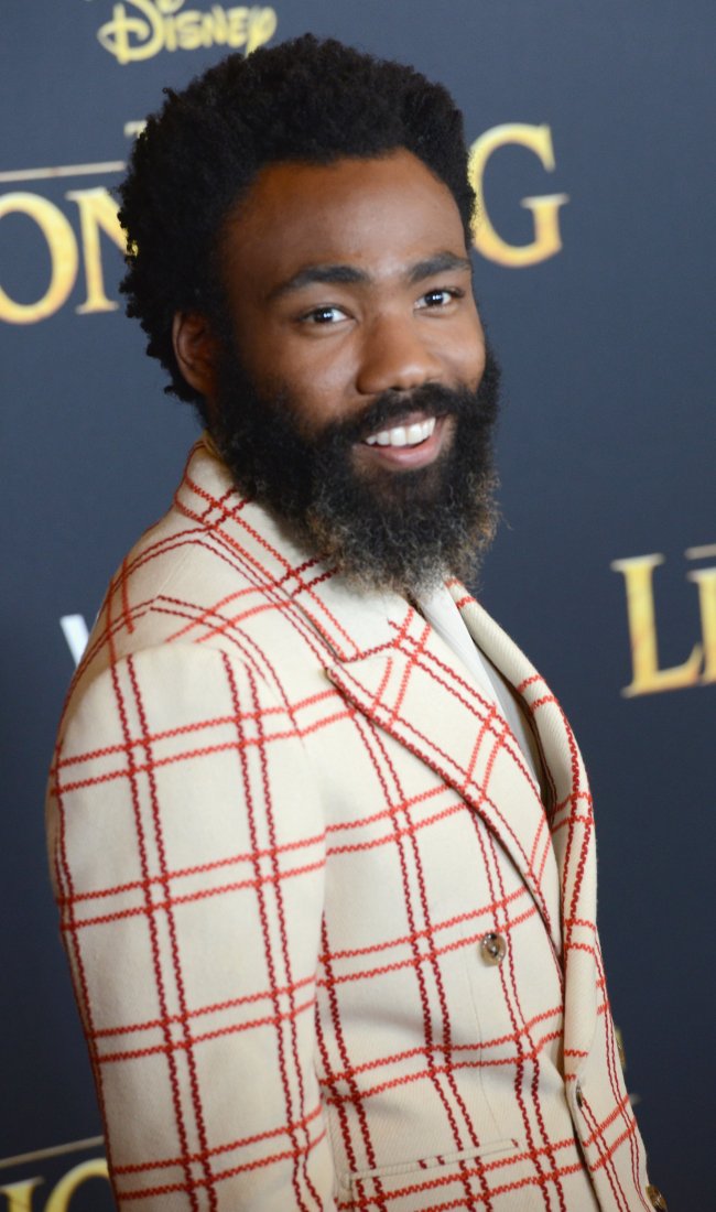 Donald Glover na premiere de O Rei Leão; ele está posando sorrindo de lado no carpet e usa um paletó branco com detalhes vermelhos quadriculados