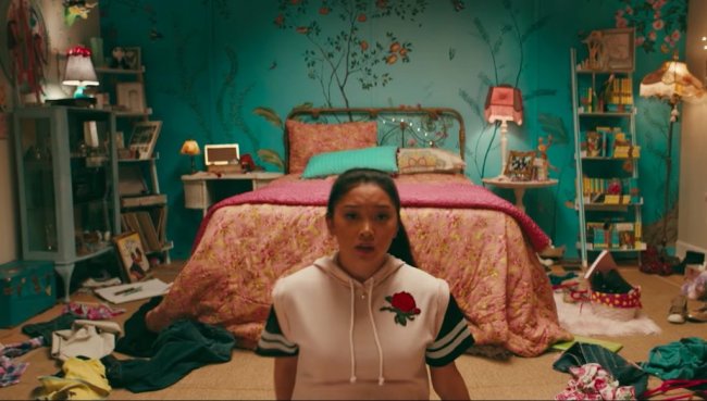 Quarto da Lara Jean de Para Todos os Garotos Que Já Amei; ela está no centro da cena com expressão assustada sentada no chão na frente da cama; a parede é decorada com flores e algumas roupas estão espalhadas pelo chão; ela usa uma blusa rosa com uma rosa bordada do lado esquerdo superior próxima ao coração; a roupa de cama é rosa com uma almofada verde turquesa; a parede é também verde turquesa com detalhes cor-de-rosa