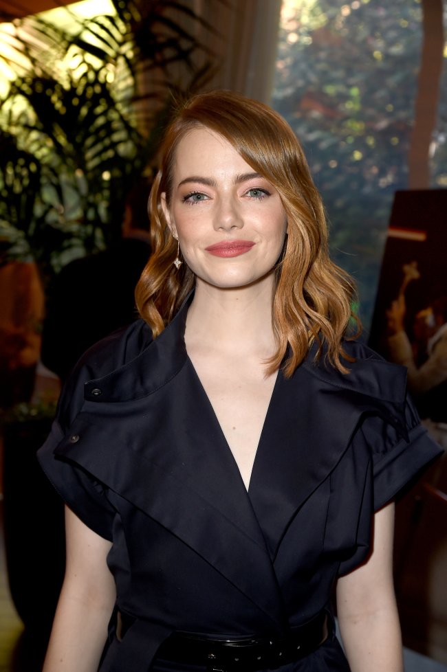 Emma Stone posando para foto; ela está olhando para câmera de frente com o cabelo solto, roupa preta de decote V com cinto da mesma cor e sorri levemente