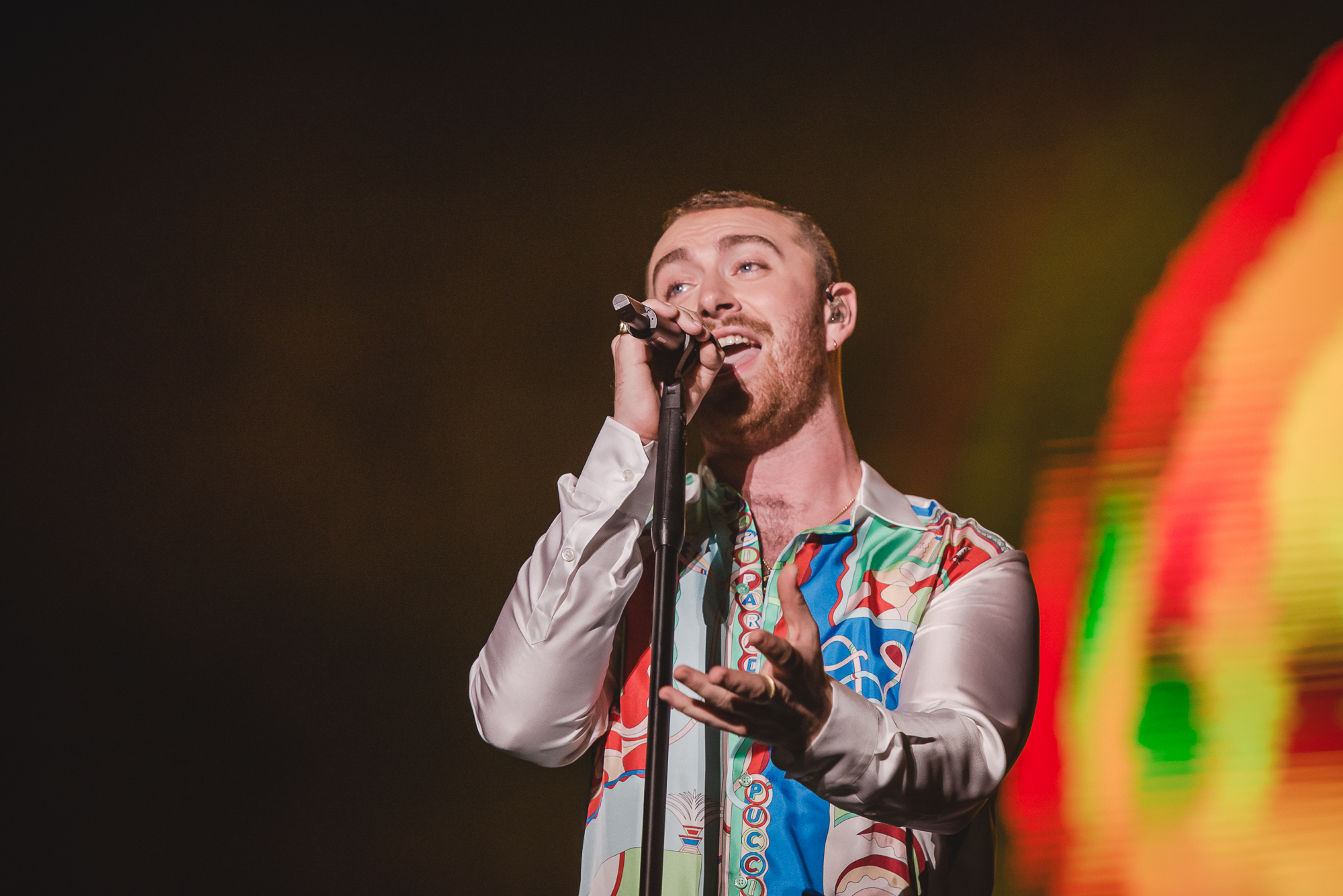 sam-smith-lollapalooza-2019 Sam Smith no palco do Lollapalooza Brasil; o cantor segura a base do microfone com uma mão e estende a outra na direção do público; ele olha para frente e luzes coloridas iluminam o fundo do palco