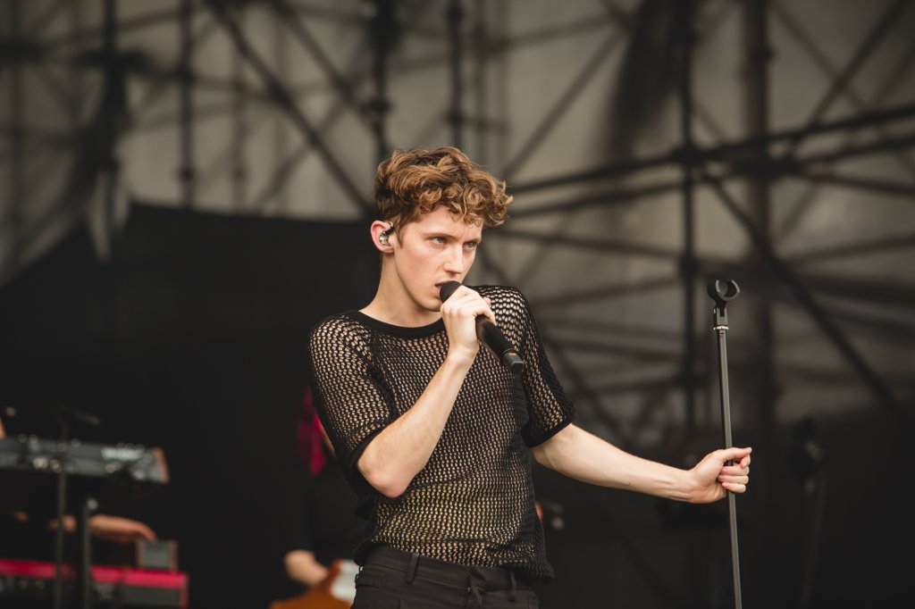 Troye Sivan cantando no palco do Lollapalooza Brasil; ele usa uma camisa preta transparente com furinhos e está observando o público enquanto canta segurando o microfone próximo do rosto