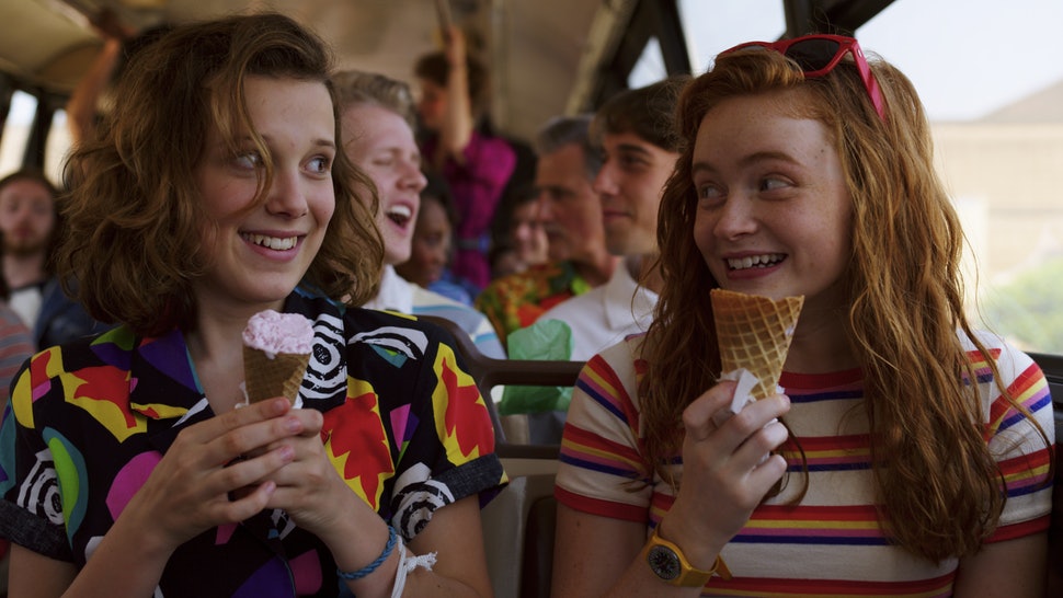 Millie Bobby Brown e Sadie Sink em cena de Stranger Things; elas estão sentadas em um ônibus olhando uma para outra segurandos sorvete e sorrindo