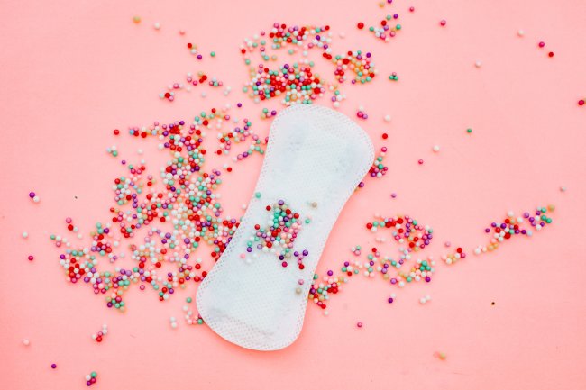 27 dúvidas esclarecidas sobre menstruação e TPM Absorvente externo sobre um fundo rosa e cheio de confeitos coloridos em cima