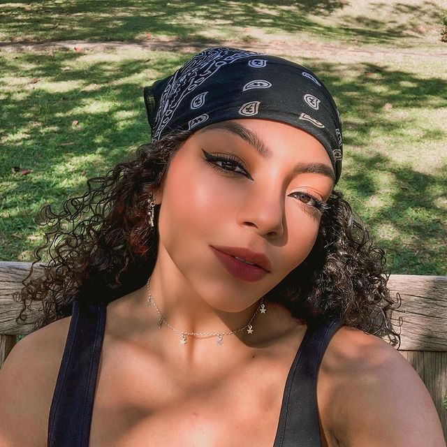 any-gabrielly-penteado-bandana Selfie da cantora Any Gabrielly. Ela usa uma regata preta, colar prata com pingentes de estrelinhas, brinco prata, maquiagem com delineador gatinho preto e batom, cabelo solto com uma bandana. Ela olha para a câmera e não sorri.