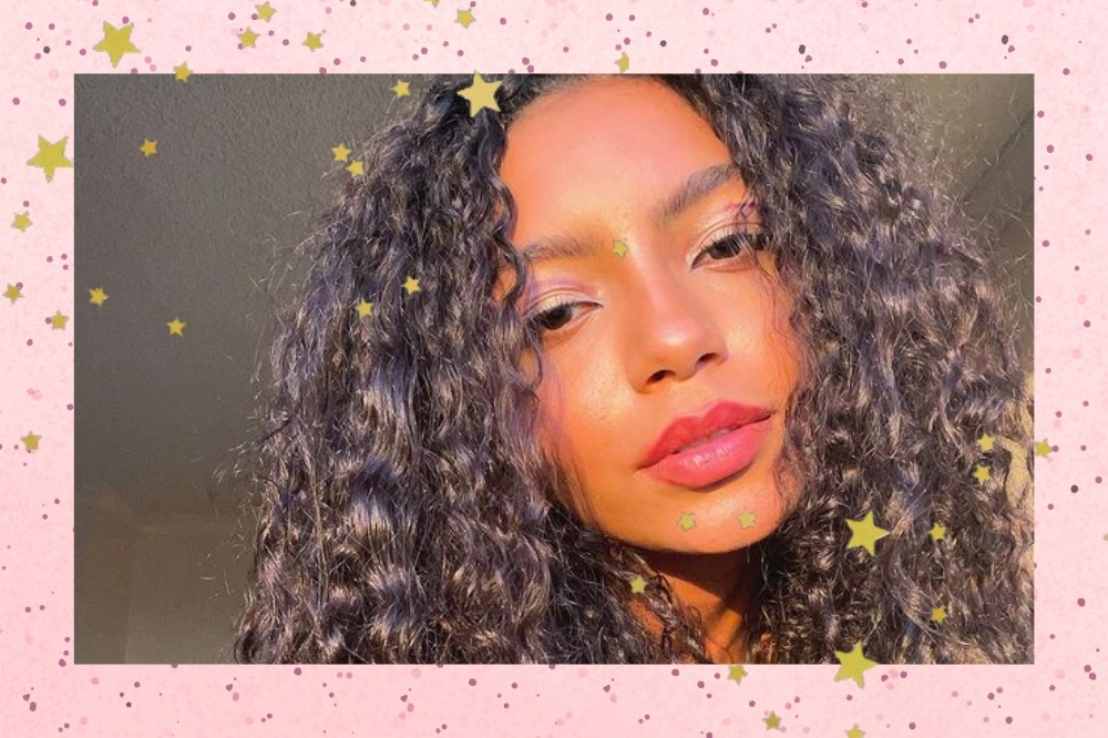 Montagem com o fundo rosa e detalhes de estrelas douradas de uma selfie da integrante brasileira do Now United, Any Gabrielly. A foto é de um close no rosto dela, ela usa uma make natural, cabelo solto, olha para a câmera e não sorri.