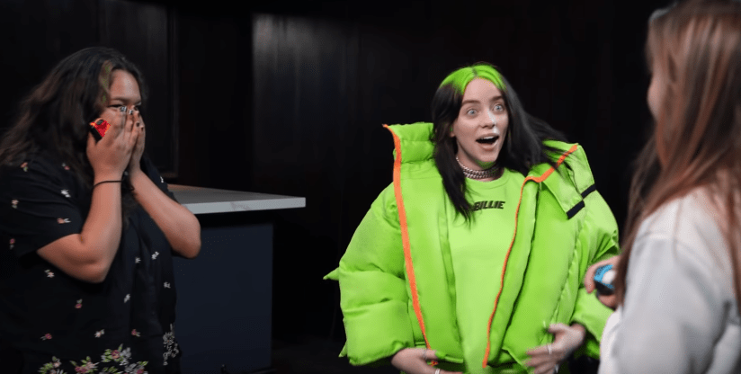 Billie Eilish surpreende fãs que testavam o Just Dance 2020; ela está com expressão surpesa ao lado de uma fã com a mão no rosto; as duas estão olhando para outra fã na frente delas