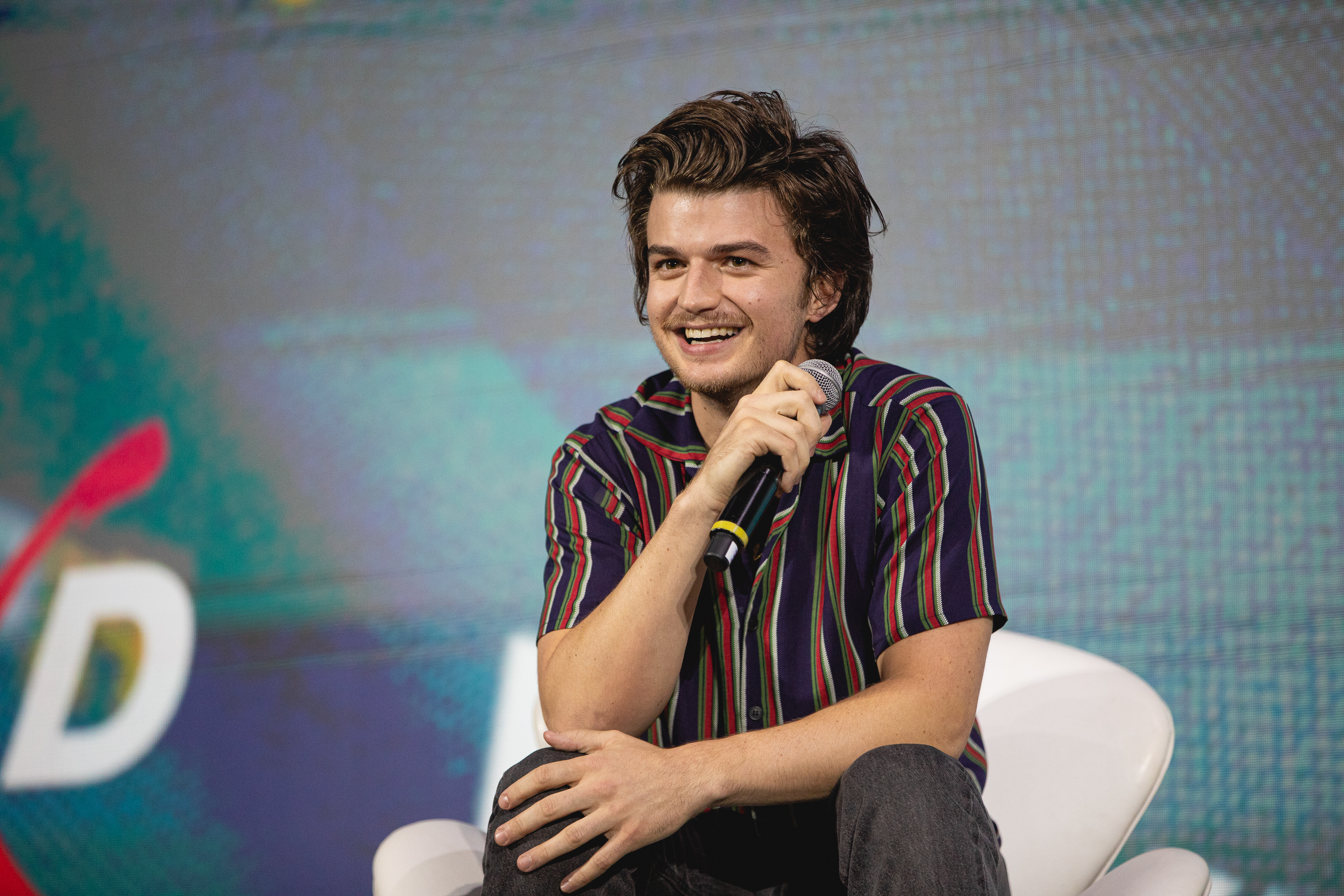 CCXP2019_Dia3_cinemark_freeguy_diegopadilha_ihf-9708 Joe Keery sentado em cadeira branca no palco do painel de Free Guy, na CCXP 2019, em São Paulo; o ator segura o microfone com uma das mãos enquanto a outra repousa em seu joelho, ele sorri observando o público e usa uma camisa listrada em tons de azul escuro, vermelho e verde