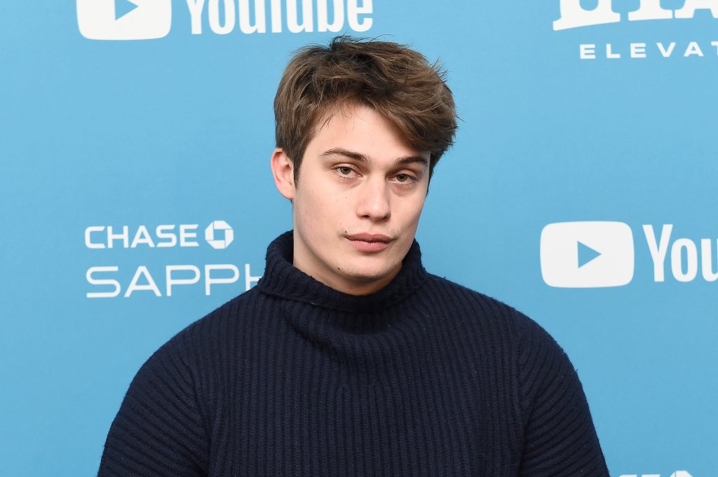 Nicholas Galitzine posando para foto; ele usa uma blusa de gola alta e está sorrindo levemente com um backdrop azul ao fundo