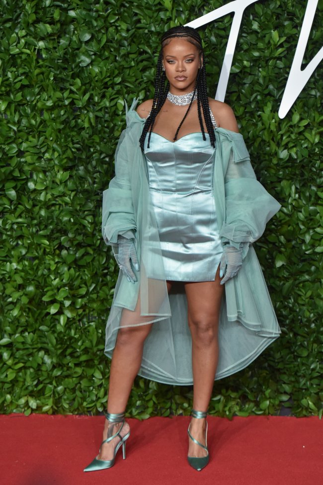 Rihanna Rihanna posando com trança, vestido azul e casado ca mesma cor. Em tapete vermelho com fundo verde. Sua expressão é séria.