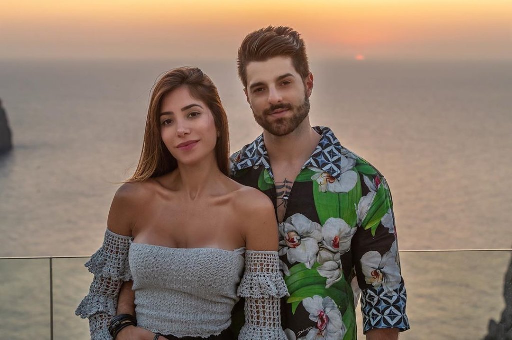 Foto de Romana e Alok; ela usa uma blusa off shoulder branca com babados na manga; ele usa uma camisa estampad em tpns de verde, preto e branco e está com a mão na cintura da médica; os dois sorriem levemente