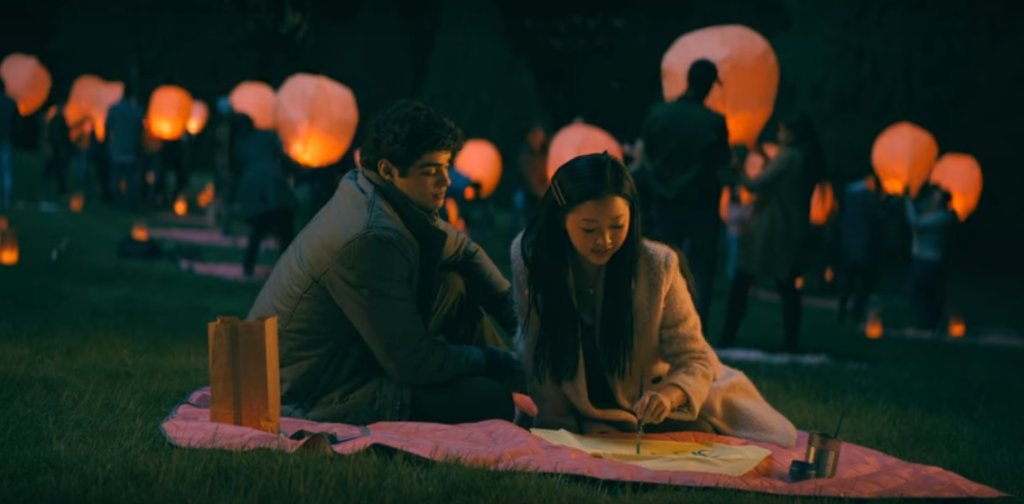 Peter K. e Lara Jean, de Para Todos os Garotos que Já Amei, sentados em uma toalha na grama durante a noite com luzes de balões ao fundo