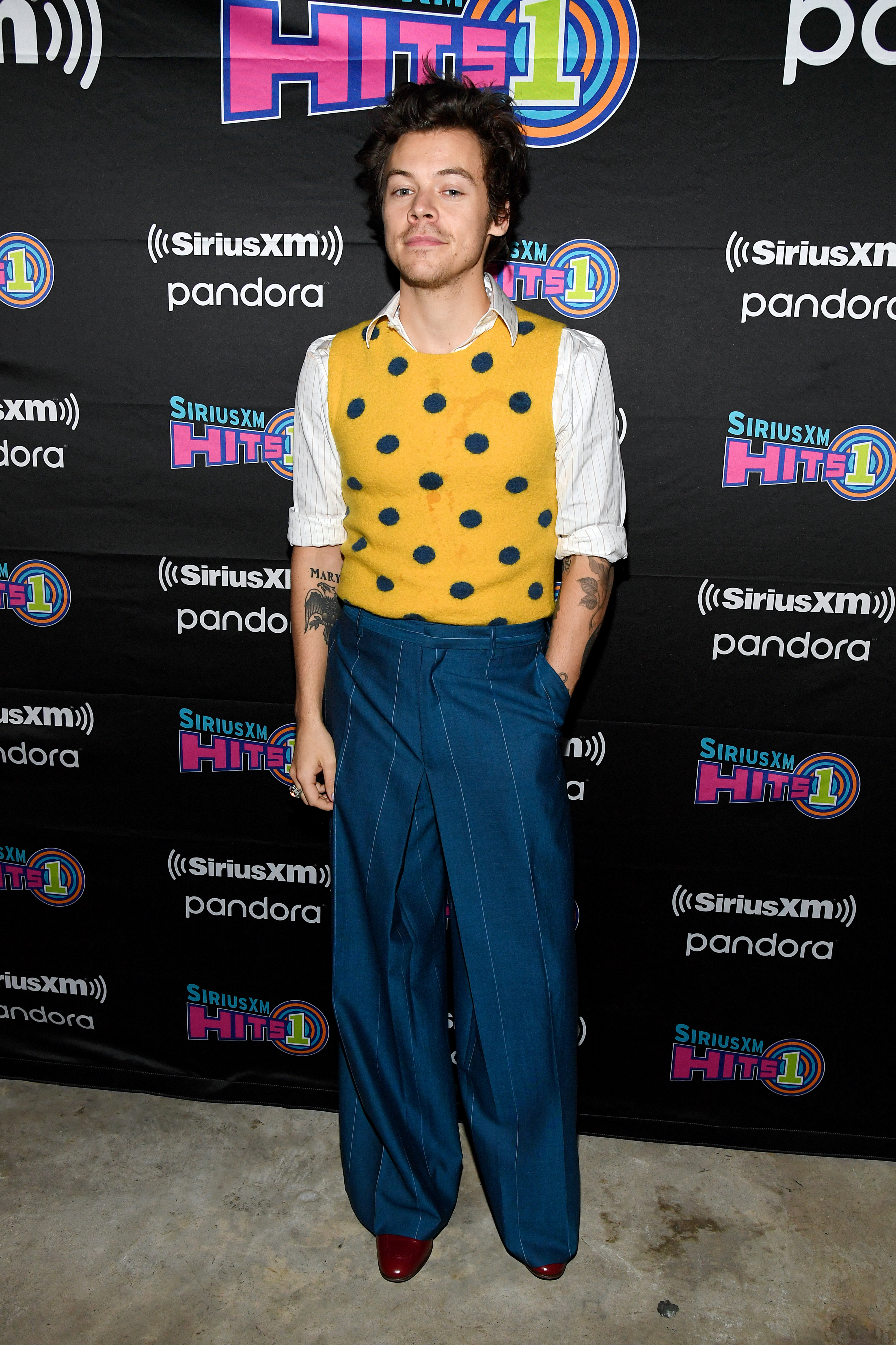 Harry Styles usando colete amarelo por cima de camisa branca com calça azul pantalona. Ele está com uma das mãos no bolso e expressão facial séria