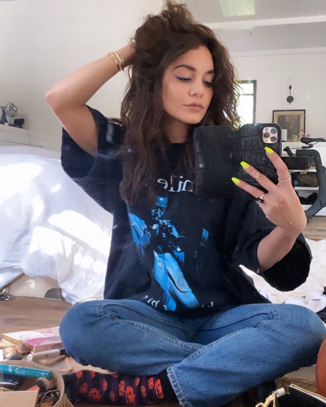 look-vanessa-hudgens-vestido A atriz aparece sentada no chão, com calça jeans, blusa preta com estampa azul, unhas amarelas e mão no cabelo posando em frente ao espelho.
