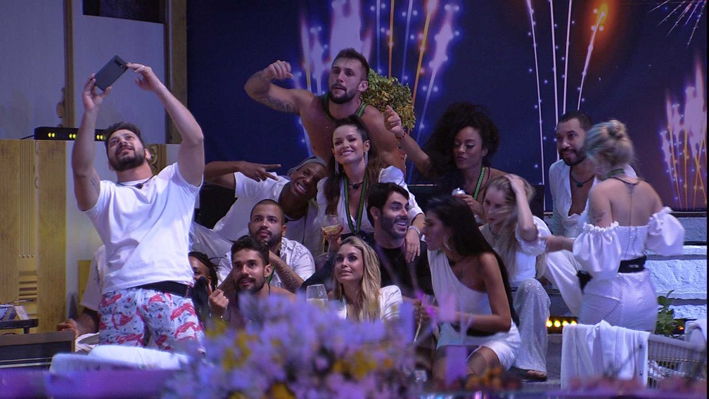 Os 20 participantes do BBB21 vestidos de branco durante uma festa do programa, eles estão reunidos para uma selfie que está sendo tirada por Caio