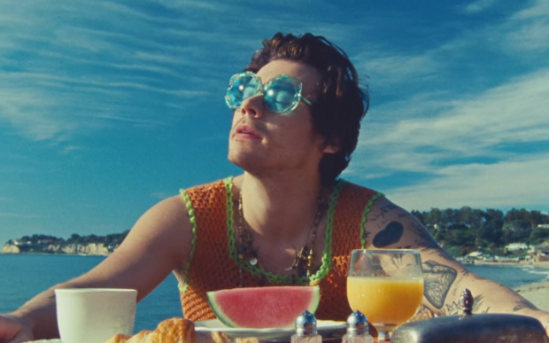 Harry Styles no clipe de Watermelon Sugar; ele está olhando para cima com óculos de sol de lente azul em uma área aberta em um dia ensolarado; na mesa ele tem uma fatia de melancia no centro com um copo de suco de laranja e uma xícara branca de cada lado da fruta; ele usa um colete de tricô laranja e verde