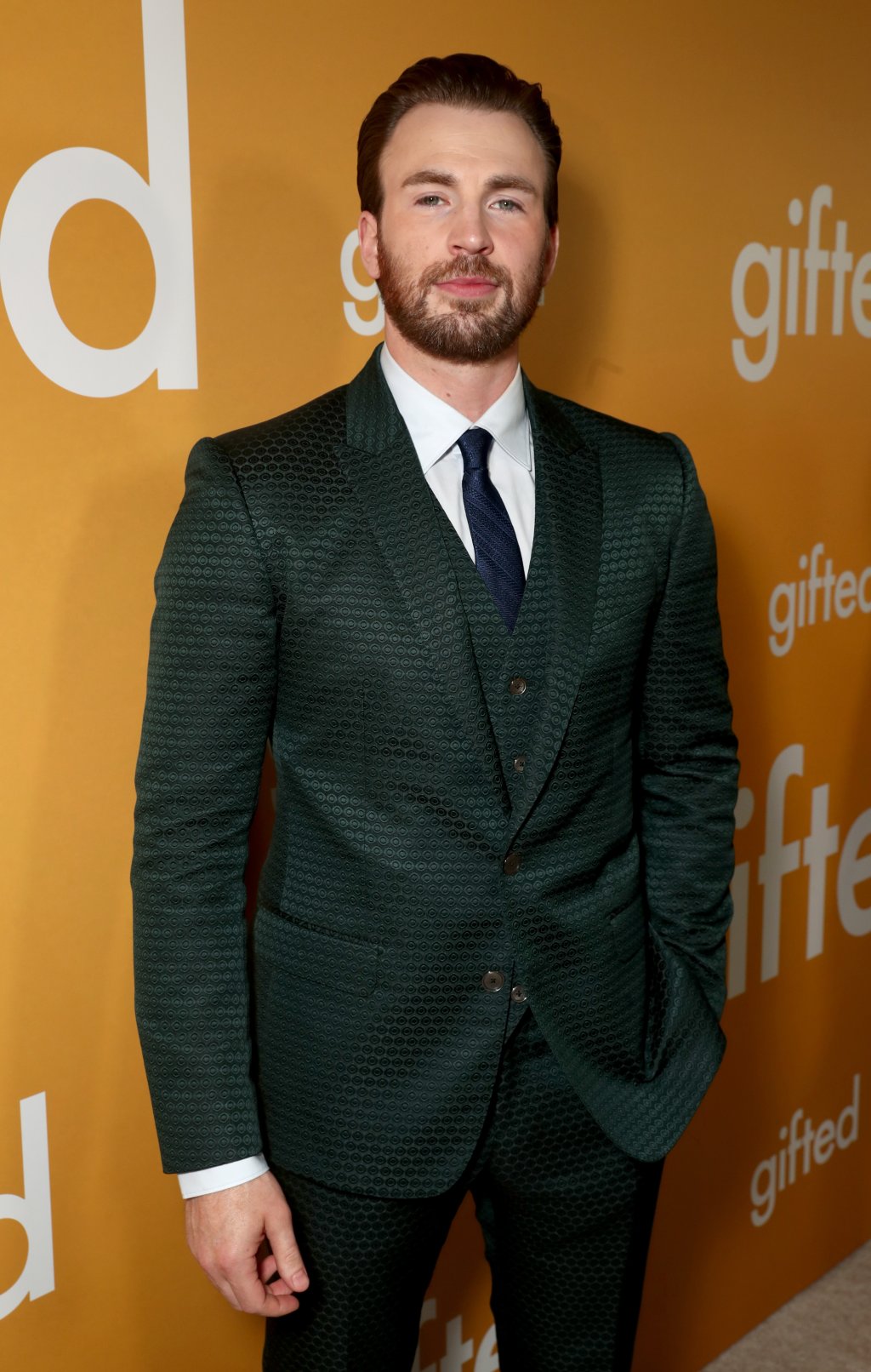 Chris Evans posando para foto em carpet com terno em tom de verde escuro e camisa branca; o ator sorri levemente com uma das mãos no bolso