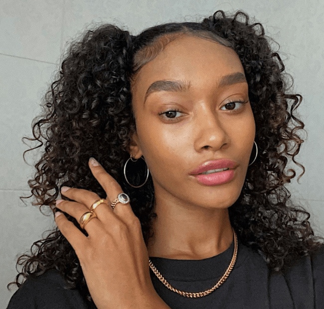 Maria-chiquinha Selfie de uma mulher. Ela usa uma camiseta preta, colar dourado, brinco de argola prateado, make natural e cabelo meio preso em uma maria chiquinha. Ela está segurando o cabelo levemente com a mão esquerda, olha para a câmera e não sorri.