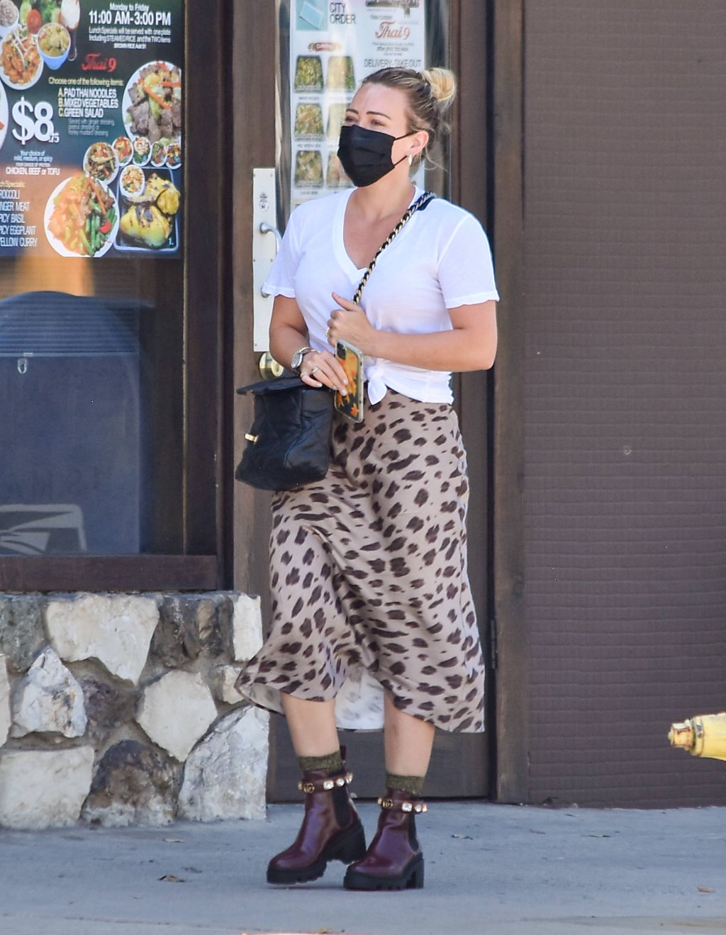 Hilary Duff usando camiseta branca, bolsa preta atravessada, saia midi com estampa de oncinha e bota marrom. Ela está com máscara de proteção preta e as duas mãos segurando a alça da bolsa enquanto anda.