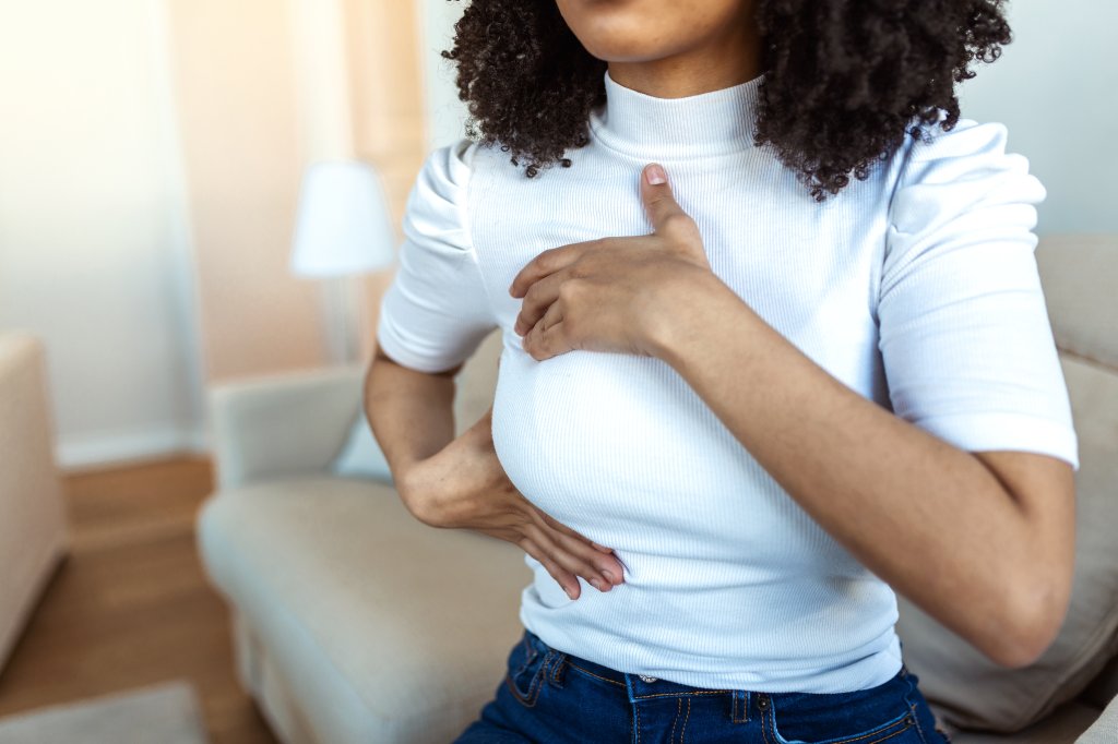 Mulher apalpando os seios. Ela usa uma blusa branca e é uma mulher negra. Não dá para ver seu rosto.