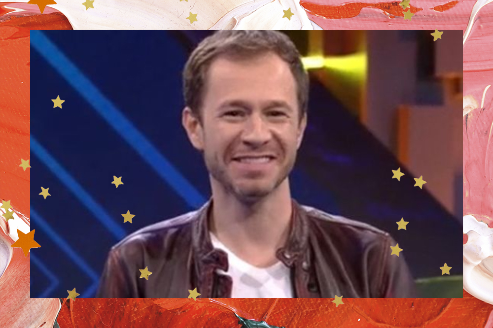 Tiago Leifert apresentando o BBB21, ele está sorrindo com camiseta branca e casaco marrom; a margem e uma textura de tintas em tons de vermelho, rosa e vermelho com estrelas amarelas decorando a imagem