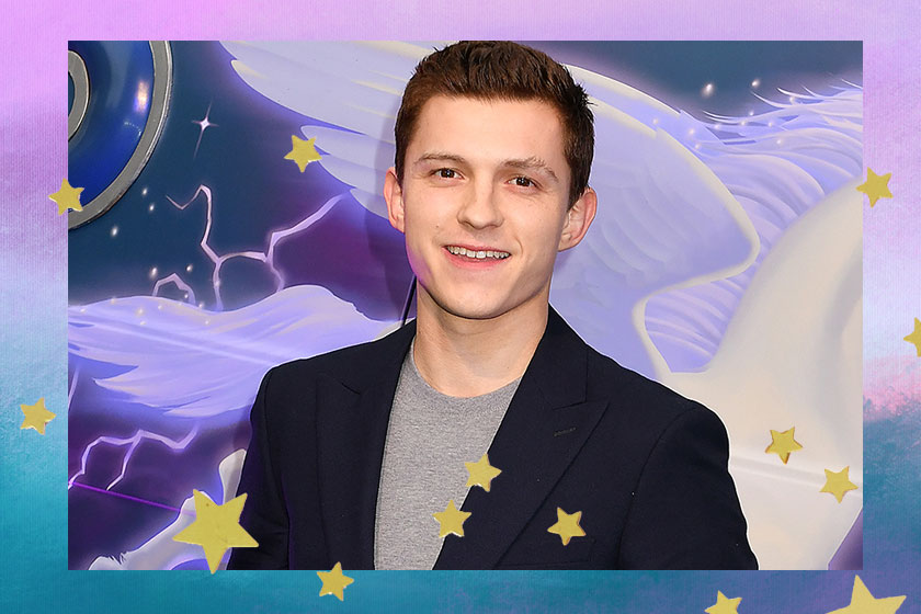 Tom Holland usando blazer azul escuro e camiseta cinza, sorrindo para a câmera