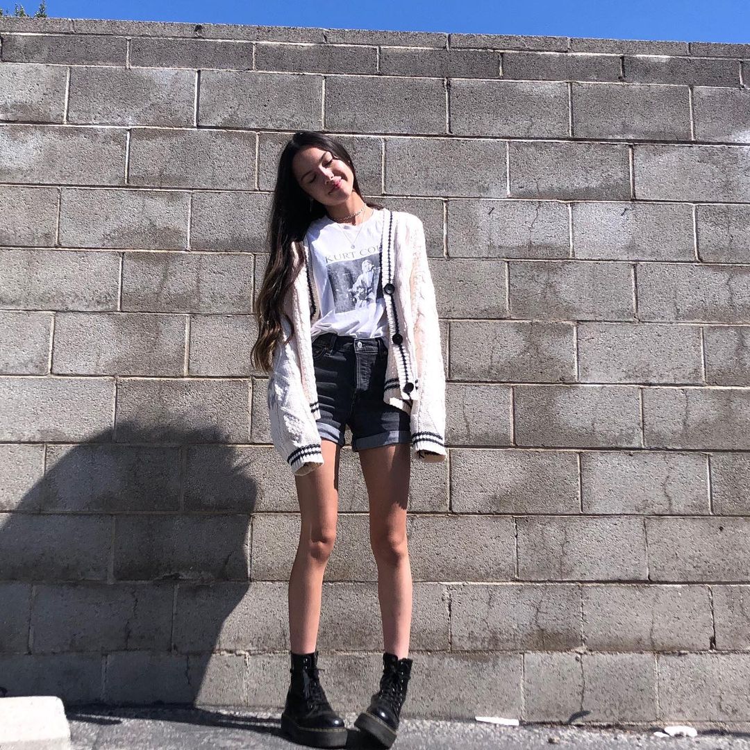 Olivia Rodrigo usando short e coturno pretos, camiseta branca e casaco bege