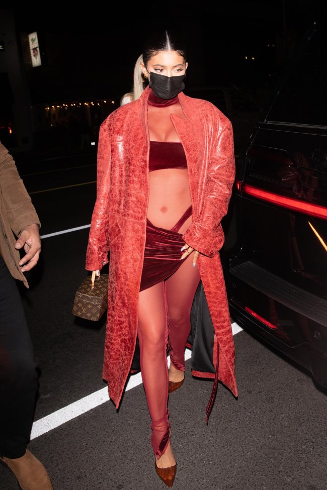 Kylie Jenner de máscara no rosto, usando macacão vermelho e transparente, com alça da calcinha à mostra com um casaco por cima