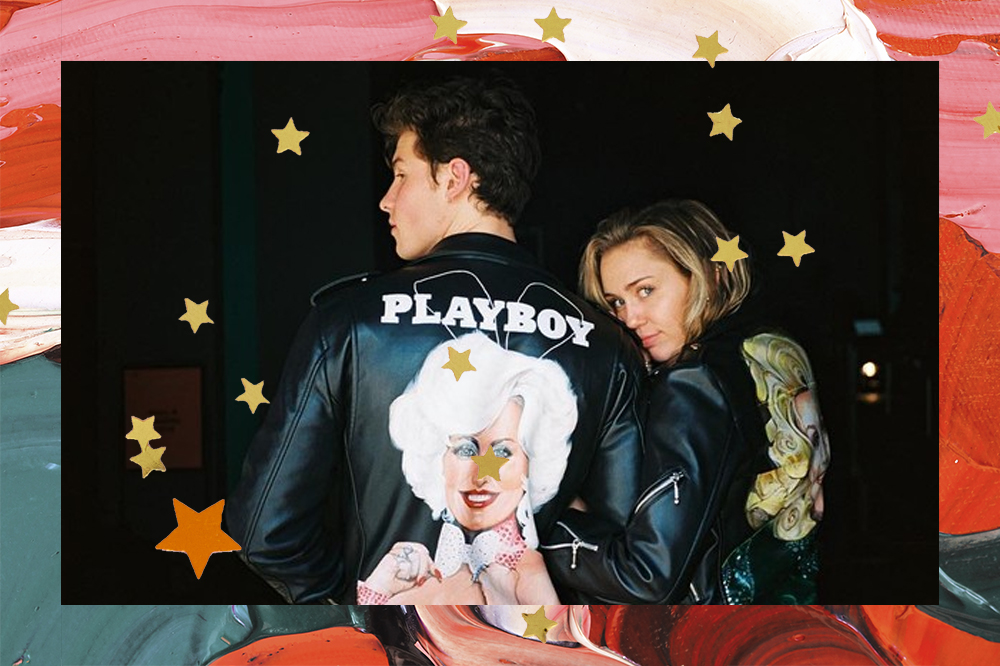 Shawn Mendes e Miley Cyrus de braços dados e de costas. Ela olha para a câmera por cima dos ombros