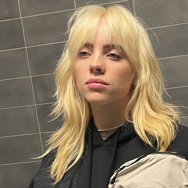 Billie Eilish Cantora Billie Eilish com corte Shaggy Hair e expressão séria, encostada na parede, usando moletom preto, atrás dela podemos observar azulejos cinza.