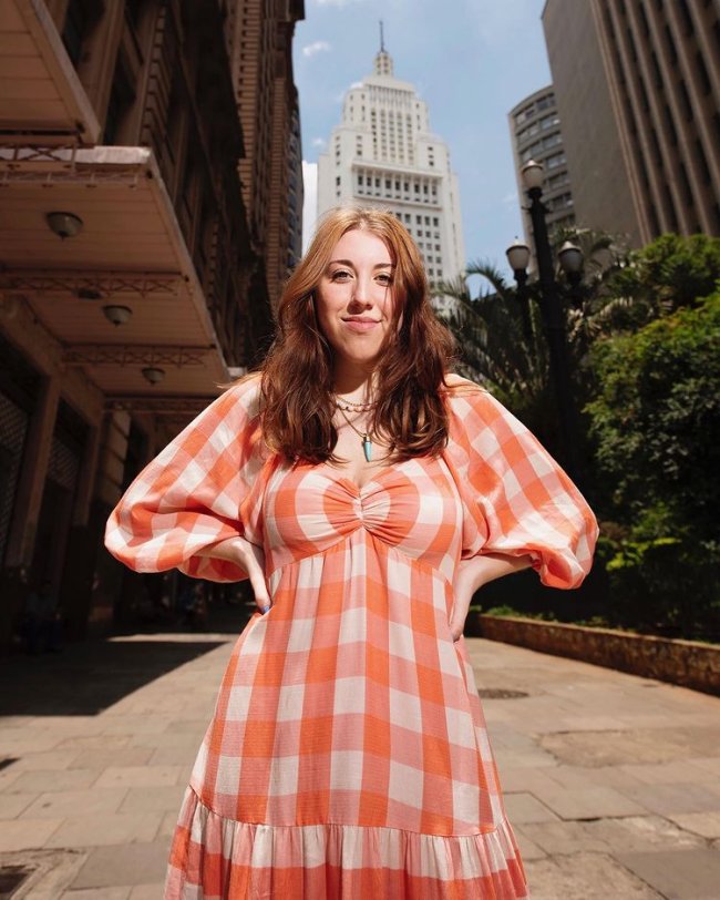 Jovem com as duas mãos na cintura, posando para foto usando vestido xadrez laranja e branco .