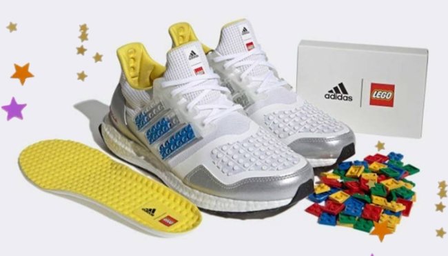 Foto com fundo branco, mostrando um par de tênis da adidas, com peças de lego espalhadas na frente do tênis, os tênis no lugar das listras pretas tradicionais da adidas, tem peça de lego azul.