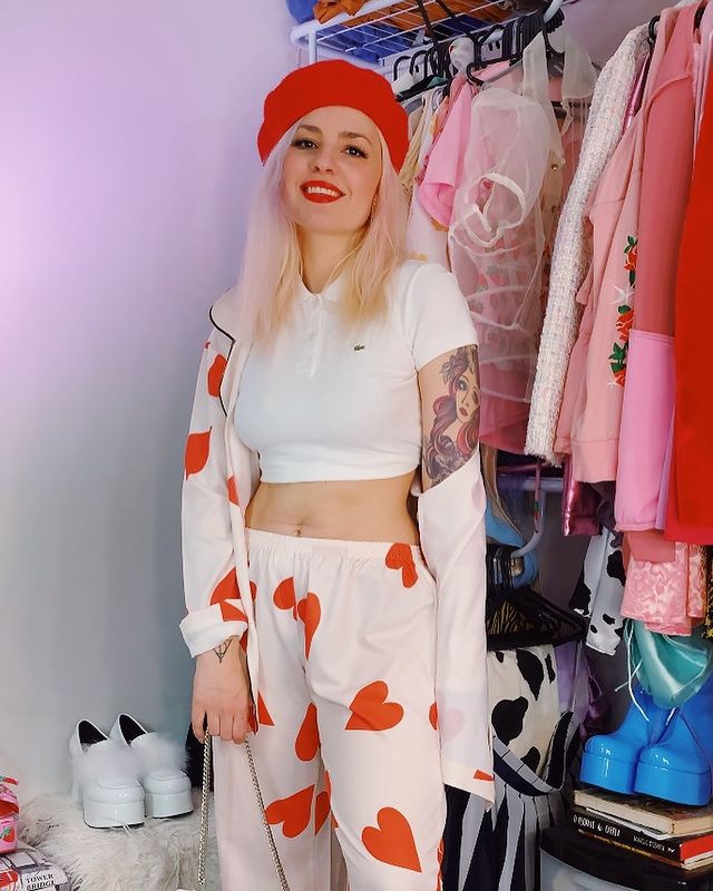 Look com uma pegada lovecore Jovem posa para foto com conjunto branco com corações rosa, conjunto composto por camisa de botões aberta e calça. Além disso, ela usa uma camiseta cropped branca, boina vermelha, e batom vermelho. Ela posa em frente a sua arara de roupas com as mais variadas peças. Sua expressão é de felicidade.