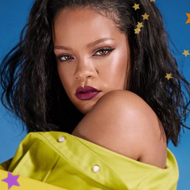 Cantora Rihanna posando para foto com delineado gatinho, batom vermelho fechado, com expressão séria, de lado, com a camisa verde caída em seu ombro.