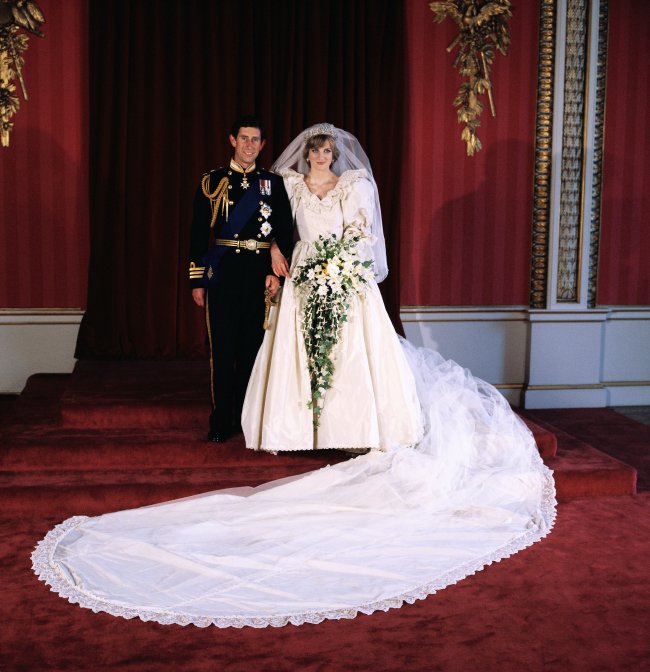 Princesa Diana e príncipe Charles em seu casamento em 1981, os dois aparecem sorridentes posando para foto. Charles usa seu traje de principe e Diana está com seu icônico vestido branco de noiva exibindo sua enorme cauda.