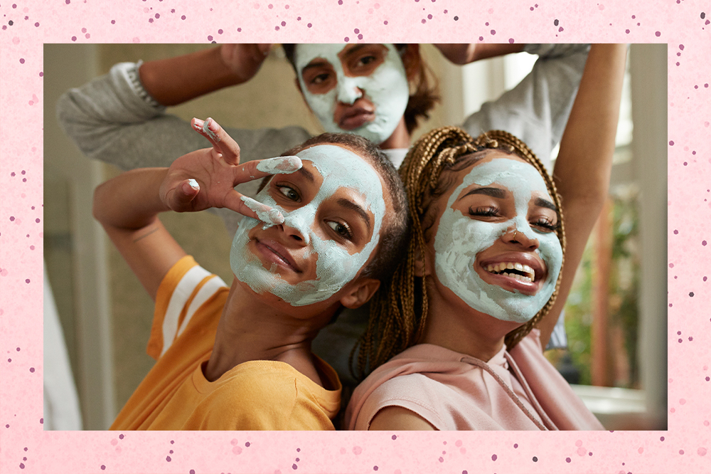 Três meninas usando máscaras faciais de argila. Duas estão na frente, uma olhando para o lado e fazendo o símbolo de paz e amor com os dedos, e a outra sorrindo com o olhar para baixo. Uma está ao fundo fazendo bico com a boca e os dois braços dobrados em direção à cabeça.