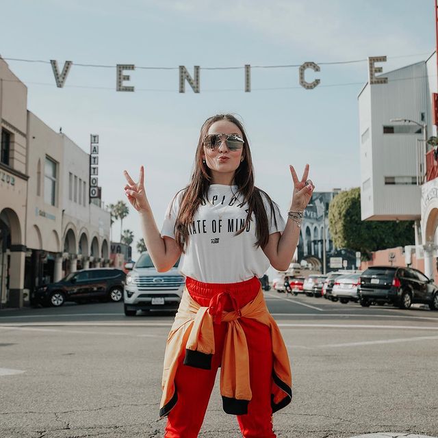 Atriz Larissa Manoela posa em rua de Venice (LA) com calça vermelha, camiseta branca e blusa laranja amarrada na cintura