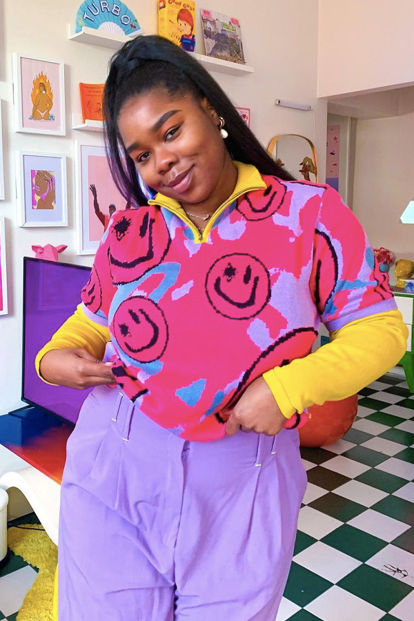 Jovem com as mãos no bolso, usando camisa com gola amarela e cor de rosa com smiles pretos, calça lilas e blusa amarela por baixo.