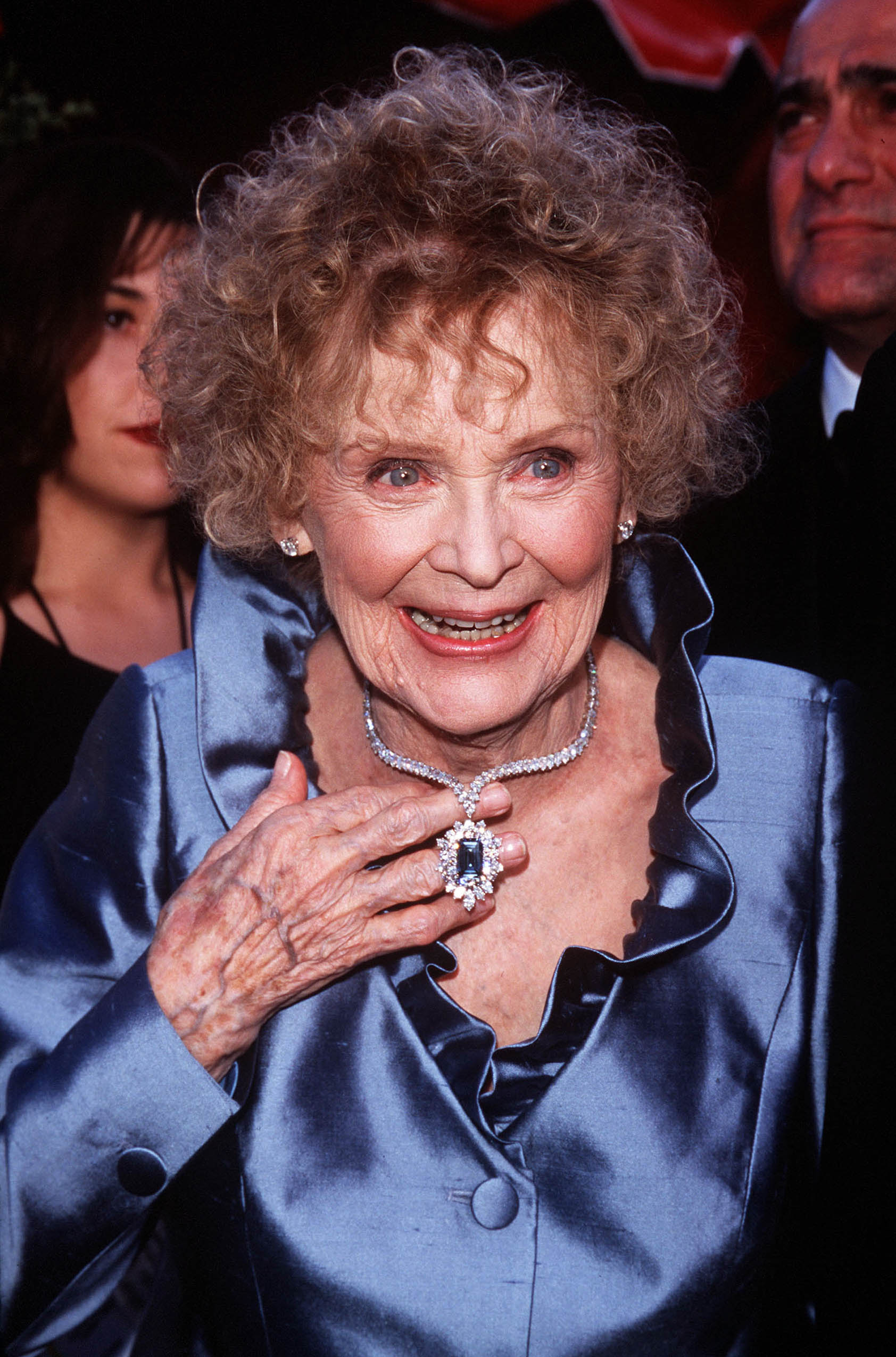 A atriz Gloria Stuart no Oscar de 1998. Ela está usando uma camisa azul de seda com babados na gola. Uma das suas mãos está segurando seu colar, que é todo de diamantes e tem um pingente com uma pedra azul no meio. Ela está sorrindo e olhando levemente para o lado direito.