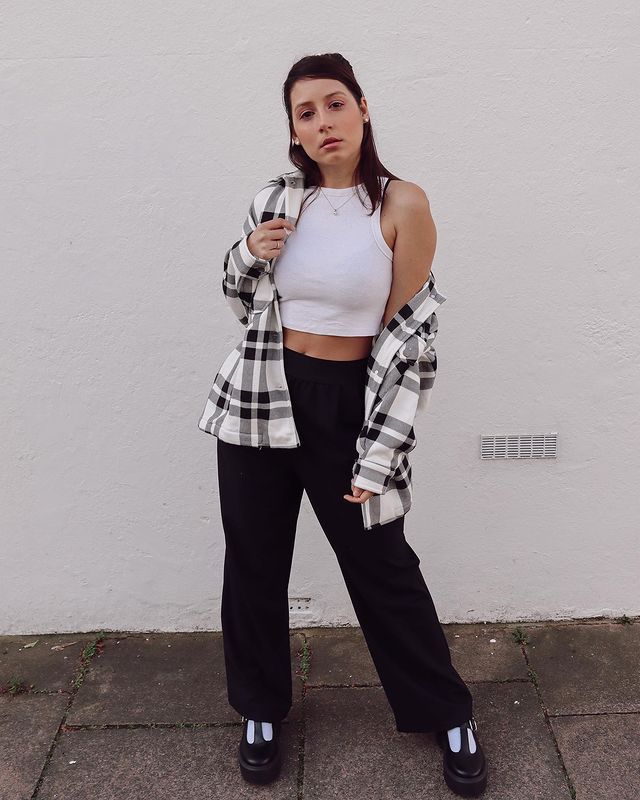 Garota usando top cropped branco com calça preta de cintura alta, sapato estilo boneca preto e meia branca. Por cima, ela usa um casaco xadrez preto e branco. Está em pé, com expressão facial séria, e uma das mãos segurando o casaco.