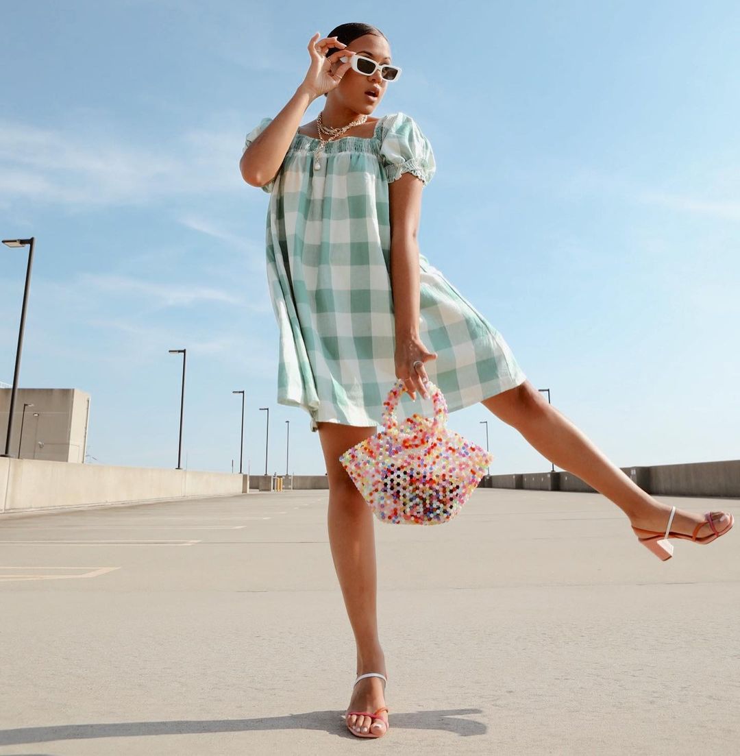 Garota usando look com vestido xadrez vichy verde e sandália branca e vermelha de tiras com saltinho. Com uma das mãos, ela segura os óculos de sol brancos, e com a outra uma bolsa de miçangas coloridas. Uma das pernas está levantada para o lado e sua expressão facial é de surpresa, com a boca aberta.