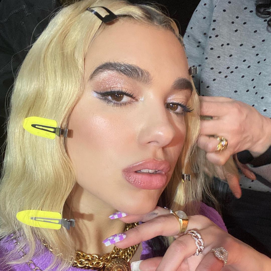 Dua Lipa usando maquiagem com sombra de brilhos e delineado fino. Ela está usando presilhas ao longo do cabelo. Está com expressão facial séria, olhando levemente para cima e uma das mãos no queixo.