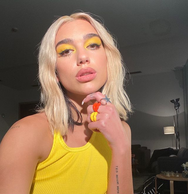 maquiagem-dua-lipa-10 Dua Lipa usando maquiagem com sombra amarela. Está usando uma regata também amarela. Uma das mãos, cheia de anéis coloridos, está tocando o queixo. Ela está com a cabeça levemente inclinada e expressão facial séria.