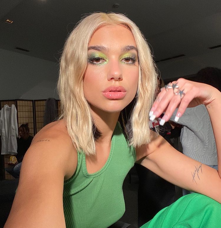 maquiagem-dua-lipa-11 Dua Lipa usando maquiagem com sombra verde com brilhos. Ela está com uma regata também verde. Um dos braços está dobrado, e suas unhas pintadas de francesinha branca. Ela está com expressão facial séria.