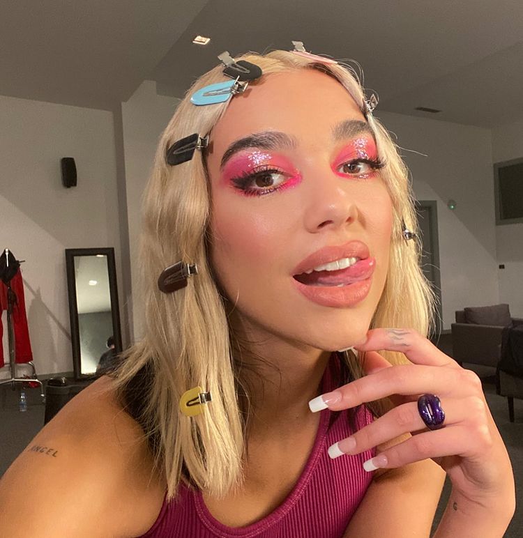 Dua Lipa usando maquiagem com sombra rosa com efeito glossy. Ela está usando uma regata no mesmo tom de rosa e presilhas ao longo da mecha frontal do cabelo. Uma das mãos está tocando o queixo. Ela está fazendo uma careta com a língua de fora.