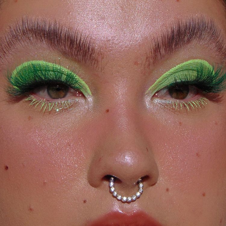 Garota usando maquiagem com dois tons de sombra verde: um mais escuro e outro mais claro. Na foto, dá apenas para ver o seu rosto. Suas sobrancelhas estão penteadas para cima, sua pele tem sardas, ela usa um piercing no septo e olha para a lateral.